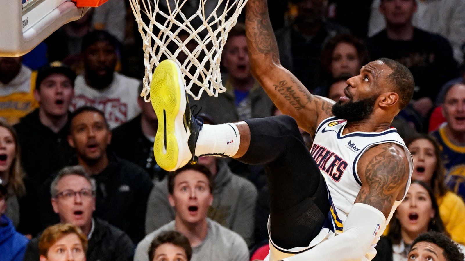 NBA比赛中一名球员飞身扣篮的定格画面，球鞋离地约一米，篮球正被强力灌入篮筐，背景是明亮的球场灯光和观众惊讶的表情